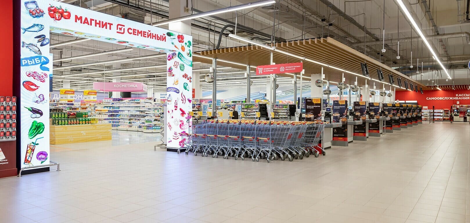 Fresh produce section in a retail superstore with overhead digital signage screens showing seasonal offers, pricing, and nutritional information